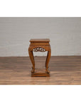 Brown wooden Ming style incense stand with cabriole legs and carved apron, placed next to a white brick wall indoors.