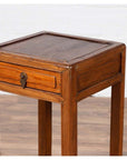 Brown elmwood lamp table with single drawer and cracked ice shelf, indoors against white wall and wood floor.