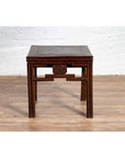 Dark wood Qing style side table with carved medallions on a wood floor next to a white brick wall.