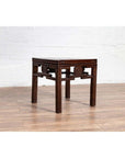 Dark wood Chinese Qing style side table with carved medallions, on wooden floor against white brick wall indoors.