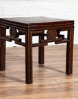 Dark wood Chinese Qing style side table with carved medallions on wooden floor next to white brick wall indoors.