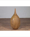 Mustard glazed ceramic vase with tapered spout on wooden floor, white brick wall in background.