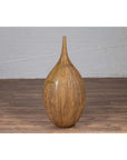 Mustard glazed ceramic vase with tapered spout on wooden floor, white brick wall in background.