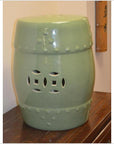 Green crackle celadon porcelain garden seat with cut-out details, placed on wood near a beige wall.
