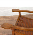 Brown wood lounge chair with curved seat and slats on a wooden floor next to a white wall indoors.