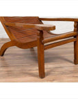 Wooden Dutch Colonial Indonesian lounge chair with curved seat and slats on a wooden floor near a white brick wall.