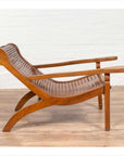 Brown wood plantation lounge chair with curved slatted seat, side view, on wooden floor next to white brick wall indoors.