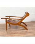 Brown wood lounge chair with curving seat and slats, on wooden floor next to white brick wall indoors.