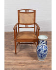 Brown teak armchair with rattan seat beside tall blue and white ceramic vase on wood floor, side view.