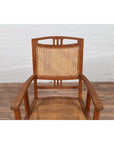 Brown teak and rattan Dutch Colonial Javanese armchair with triglyph motifs on wooden floor by white brick wall.
