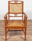 Brown teak and rattan armchair with triglyph motifs on wooden floor against a white brick wall.