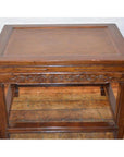 Brown elmwood stool or table with carved detailing, placed on wooden floor in front of a white brick wall.
