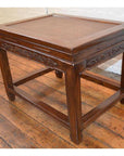 Brown elmwood stool or table with carved floral details and straight legs on wooden floor beside white brick wall.