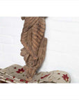 Brown wooden Indian deity statue on floral-embroidered fabric, white brick wall background, indoor setting.