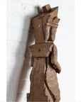 Brown hand-carved wooden statue of a Hindu deity from Gujarat, set against a white brick wall indoors.