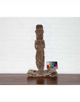 Brown wood carved statue on folded textile next to color checker, white brick wall background.