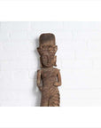 Brown wood hand-carved Hindu deity statue from Gujarat, indoors against a white brick wall.