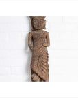 Brown wood hand carved Hindu deity statue from Gujarat, displayed indoors against a white brick wall.