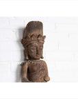 Brown hand-carved wooden statue of a Hindu deity with broken crown, set against white brick background.