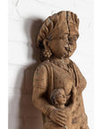 Brown wood statue of mother and child, hand carved in Indian temple style, set against a white brick wall indoors.