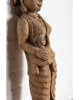 Brown wood mother and child statue, hand carved, Indian temple style, placed in front of a white brick wall indoors.