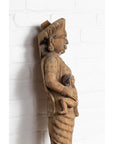 Brown wood mother and child temple carving statue, hand carved, side view, against white brick wall indoors.