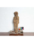 Brown hand-carved wood statue of mother and child from Gujarat, shown with color card against white brick background.