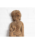 Brown wood mother and child statue, hand-carved with Indian temple design, placed in front of a white brick wall.