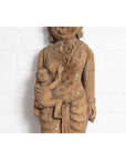 Brown hand-carved wooden temple statue of a mother holding her child, set against a white brick wall.
