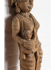 Brown wooden statue of mother and child, hand carved in Indian temple style, with white brick background.