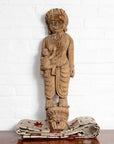 Brown wooden statue of mother and child with Indian temple carvings, displayed on patterned fabric against white brick wall.