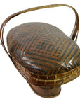 Brown rattan basket with curved handle and geometric patterned lid, hand woven, shown without background.