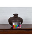 Brown metal Indian 19th c. water jar with wide belly and protruding lip on wood surface, color card in front.