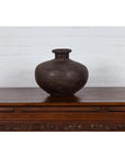 Dark clay vase and 19th century Indian metal water jar on carved table against white brick wall.