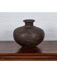 Bronze metal Indian 19th-century water jar with rounded belly and protruding lip on wood surface, white brick wall background.