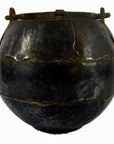Close-up of round blackened iron water jug with handle and worn finish, used as vintage vase or home decor.