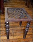 Brown wooden Indian side table with carved legs, lattice seat, and iron top on a wooden floor.