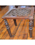 Dark wood Indian side table with ornate carving and four turned legs near window grill on polished wooden floor.