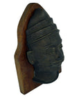 Dark brass bas-relief plaque of sculpted head with conical headdress, mustache, mounted on wood, close-up.