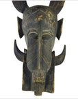 Brown wooden Indonesian hand carved figural shield with elongated features, horns, and geometric patterns on white background.