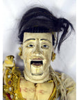 Close-up of a dark-haired Indonesian puppet with tangled strings and a moving mouth, set against a white background.