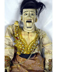 Close-up of Indonesian puppet with painted face, ornate jeweled clothing, black hair, and articulated fingers.
