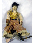 Brown wooden Indonesian puppet with detailed facial features, moving mouth, and articulated fingers, seated on plain backdrop.
