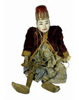 Close-up of painted Indonesian puppet with red hat, velvet jacket, and tangled limb strings, seated upright.