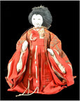 Close-up of a Japanese doll with a white face, black hair, and red-gold kimono against a black background.