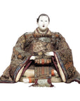 Close-up of Japanese doll in ornate Taisho Period kimono with white face and hands, seated against plain background.