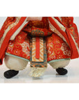 Close-up of Japanese doll in ornate red and gold fabric, kneeling with black shoes on white background.