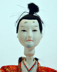 White-faced Japanese doll with black hair, red and gold clothing, on plain background.