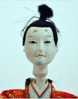 White-faced Japanese doll with black hair, red and gold clothing, on plain background.