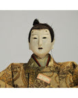 Close-up of Japanese Taisho samurai doll with white mask-like face and ornate patterned clothing, plain background.